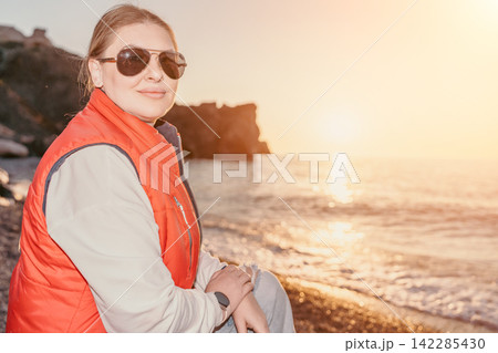 Woman beach sunset smiling woman in sunglasses and red vest sits on pebble shore by ocean at golden hour with copy space 142285430