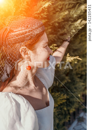 Woman forest vintage wearing a net headpiece and cherry earring stands in sunlight for a romantic lifestyle portrait Woman forest vintage wearing a net headpiece and cherry earring stands in sunlight for a romantic lifestyle portrait 142285431