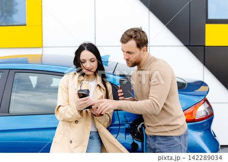 Young couple man and woman traveling by electric car having stop at charging station. Boyfriend plugging in cable to charge. Man talking with girlfriend, holding cup drinking hot coffee smiling Young couple man and woman traveling by electric car having stop at charging station. Boyfriend plugging in cable to charge. Man talking with girlfriend, holding cup drinking hot coffee smiling 142289034