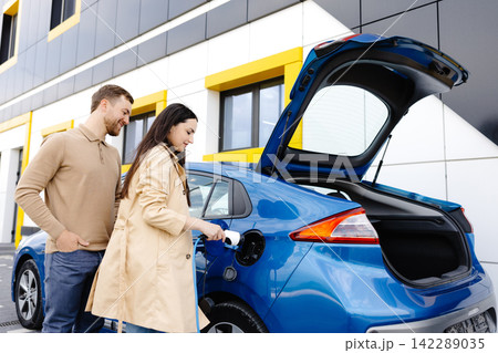 Young couple man and woman traveling by electric car having stop at charging station. Girlfriend holding charger looking at car and smiling Young couple man and woman traveling by electric car having stop at charging station. Girlfriend holding charger looking at car and smiling 142289035