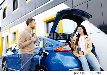Young couple man and woman traveling by electric car having stop at charging station. Boyfriend plugging in cable to charge. Man talking with girlfriend, holding cup drinking hot coffee smiling Young couple man and woman traveling by electric car having stop at charging station. Boyfriend plugging in cable to charge. Man talking with girlfriend, holding cup drinking hot coffee smiling 142289036