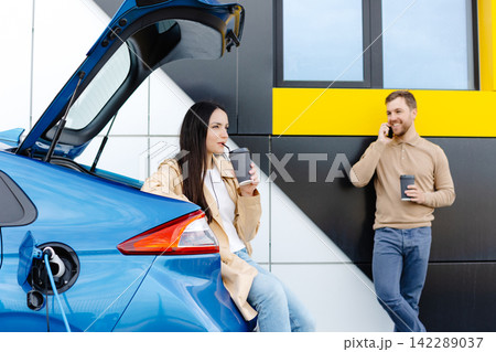 Young couple man and woman traveling together by new car having stop at charging station. Boyfriend holding cup drinking hot coffee talking to the phone and girlfriend looking aside and smiling Young couple man and woman traveling together by new car having stop at charging station. Boyfriend holding cup drinking hot coffee talking to the phone and girlfriend looking aside and smiling 142289037