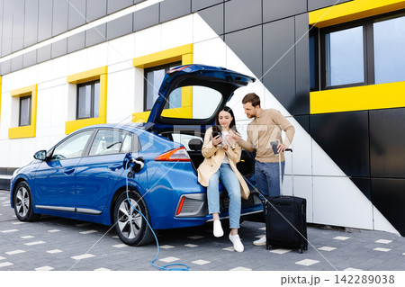 Young friends charging automobile battery from public station and standing near parking for electric car. Man and woman talking and drinking coffee in city Young friends charging automobile battery from public station and standing near parking for electric car. Man and woman talking and drinking coffee in city 142289038