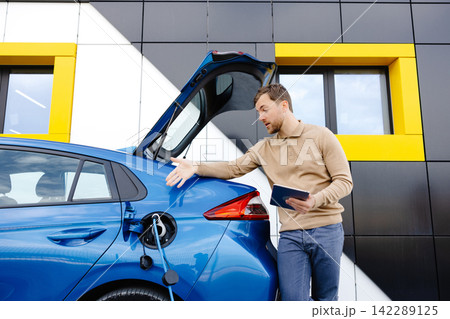 A young man is charging an electric car while standing at a station and holding a tablet A young man is charging an electric car while standing at a station and holding a tablet 142289125