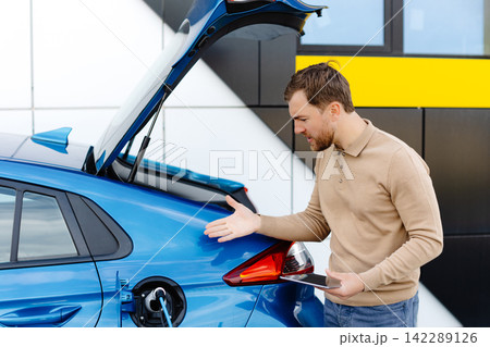 A young man is charging an electric car while standing at a station and holding a tablet A young man is charging an electric car while standing at a station and holding a tablet 142289126