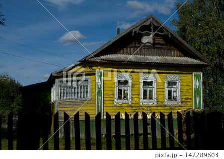 Old yellow wooden village house with carved window frames behind fence Old yellow wooden village house with carved window frames behind fence 142289603