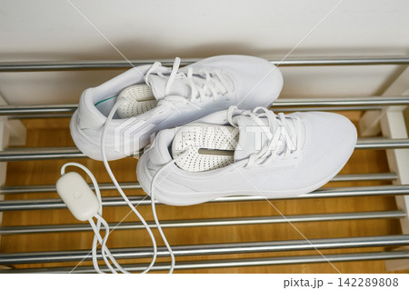 Electric shoes dryer top view. Pair of white wet sneakers drying with a plug-in boot warmer on a shoe rack. The concept of household technologies and smart gadgets. Electric shoes dryer top view. Pair of white wet sneakers drying with a plug-in boot warmer on a shoe rack. The concept of household technologies and smart gadgets. 142289808