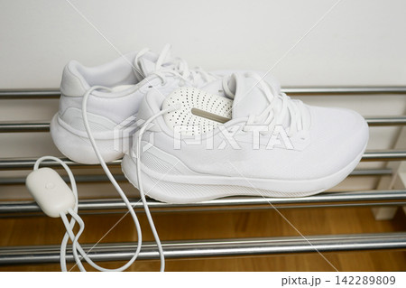 Electric shoes dryer. Pair of white wet sneakers drying with a plug-in boot warmer on a shoe rack. The concept of household technologies and smart gadgets. 142289809