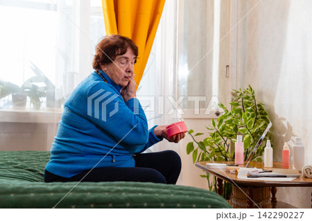 Elderly Caucasian woman applying face cream while sitting comfortably on a bed at home. The skincare routine represents self care, beauty, confidence, and a positive approach to aging. Side view Elderly Caucasian woman applying face cream while sitting comfortably on a bed at home. The skincare routine represents self care, beauty, confidence, and a positive approach to aging. Side view 142290227