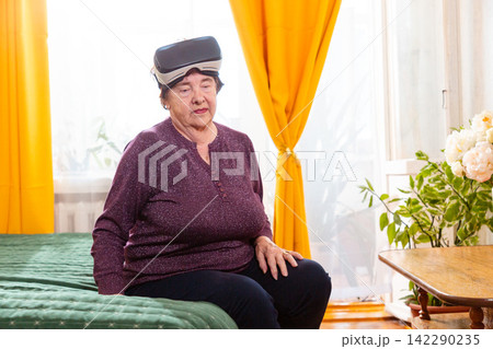 Senior Caucasian woman enjoys a virtual reality experience while sitting on her bed at home. Modern gadgets and digital entertainment support contemporary retirement. Mid shot Senior Caucasian woman enjoys a virtual reality experience while sitting on her bed at home. Modern gadgets and digital entertainment support contemporary retirement. Mid shot 142290235