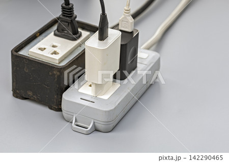 Close-up of an old, dusty electrical power strip and extension outlet overloaded with multiple phone chargers, adapters, and tangled cables all plugged in simultaneously Close-up of an old, dusty electrical power strip and extension outlet overloaded with multiple phone chargers, adapters, and tangled cables all plugged in simultaneously 142290465