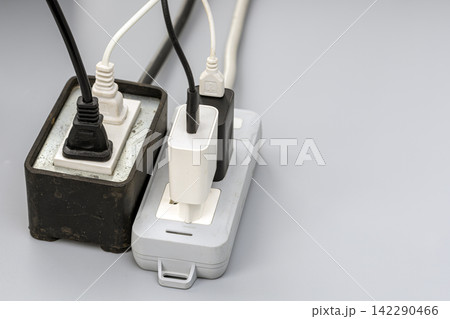 Close-up of two old, dirty power strips overloaded with multiple tangled black and white electrical cables, USB chargers, and plugs, symbolizing electrical safety hazards and overload risk Close-up of two old, dirty power strips overloaded with multiple tangled black and white electrical cables, USB chargers, and plugs, symbolizing electrical safety hazards and overload risk 142290466