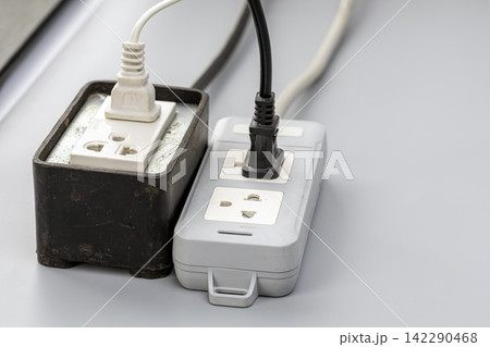 Close-up view of two old, dusty power strips with white and black electrical cords plugged into multiple outlet sockets, showing wear, dust, and daily household electrical usage Close-up view of two old, dusty power strips with white and black electrical cords plugged into multiple outlet sockets, showing wear, dust, and daily household electrical usage 142290468
