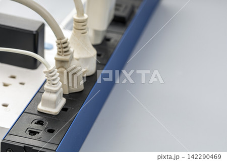 Detailed close-up view of several white electrical plugs connected to a black and blue power strip surge protector, showing multiple outlets used simultaneously on an office desk Detailed close-up view of several white electrical plugs connected to a black and blue power strip surge protector, showing multiple outlets used simultaneously on an office desk 142290469