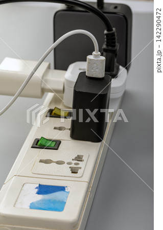 Detailed close-up of a white electrical power strip overloaded with multiple plugs, black and white adapters, colored indicator switches, and a white USB charging cable snaking upward Detailed close-up of a white electrical power strip overloaded with multiple plugs, black and white adapters, colored indicator switches, and a white USB charging cable snaking upward 142290472
