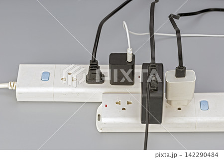 Close-up view of two white electrical power strips crowded with multiple black and white power cables, USB chargers, and adapter plugs, illustrating electrical overload and cluttered wiring against Close-up view of two white electrical power strips crowded with multiple black and white power cables, USB chargers, and adapter plugs, illustrating electrical overload and cluttered wiring against 142290484