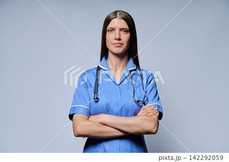 Confident female nurse in blue uniform with stethoscope standing with arms crossed against grey background Confident female nurse in blue uniform with stethoscope standing with arms crossed against grey background 142292059