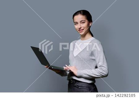 Young female student holding laptop computer and looking at camera on grey background Young female student holding laptop computer and looking at camera on grey background 142292105