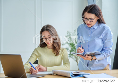Female teacher supervising young student writing notes at desk with laptop Female teacher supervising young student writing notes at desk with laptop 142292127