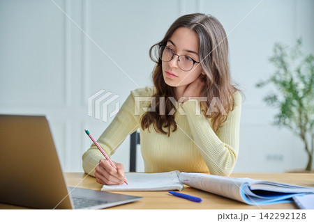 Focused female student taking notes at desk with laptop front view Focused female student taking notes at desk with laptop front view 142292128