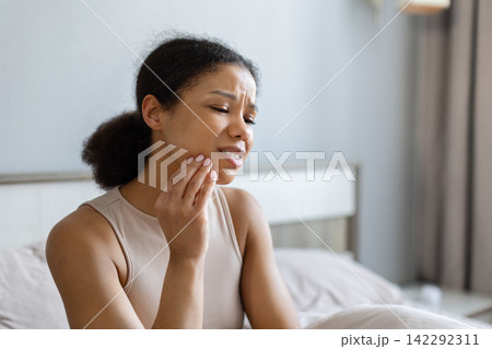 Young Black woman in bedroom touching jaw with painful expression, experiencing toothache Young Black woman in bedroom touching jaw with painful expression, experiencing toothache 142292311