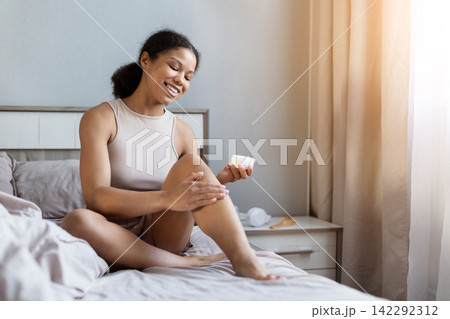 Smiling young Black woman sitting on bed applying moisturizing cream to leg Smiling young Black woman sitting on bed applying moisturizing cream to leg 142292312