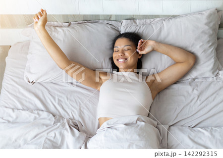 Smiling Black woman stretching her arms while lying in bed after waking 142292315
