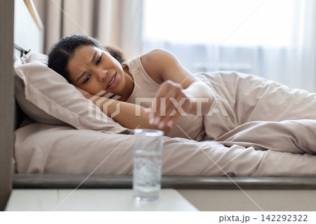 Young Black woman lying in bed reaching toward glass of water Young Black woman lying in bed reaching toward glass of water 142292322