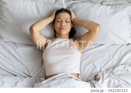 Young Black woman lies in bed holding head, experiencing headache or insomnia Young Black woman lies in bed holding head, experiencing headache or insomnia 142292323