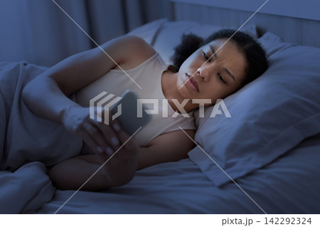 Worried Black woman lying awake in bed at night checking her smartphone Worried Black woman lying awake in bed at night checking her smartphone 142292324