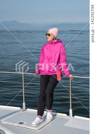 Woman in pink jacket and beanie stands on catamaran bow against Pacific Ocean and Kamchatka mountains. Marine travel, adventure tourism, vacation, freedom, outdoor lifestyle. 142292598