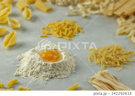 Variety of uncooked pasta with flour and egg yolk forming a circle on a gray background Variety of uncooked pasta with flour and egg yolk forming a circle on a gray background 142292618