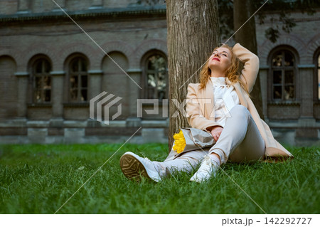 Woman feeling relaxed while sitting on green grass, leaning against a tree trunk, enjoying the sun and fresh air with closed eyes, holding yellow flowers Woman feeling relaxed while sitting on green grass, leaning against a tree trunk, enjoying the sun and fresh air with closed eyes, holding yellow flowers 142292727