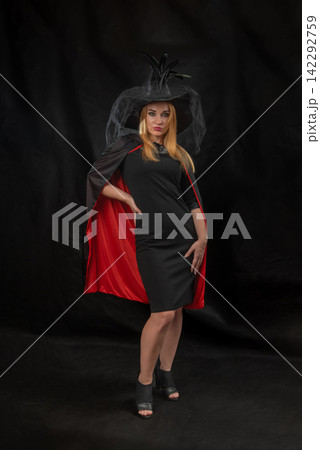 Woman standing confidently in a black dress, a red lined black cape, and a witch hat with feathers and a veil, celebrating Halloween or a costume party 142292759