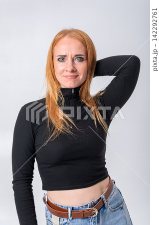 Young woman with red hair and green eyes posing in a studio, displaying a thoughtful expression with slight uncertainty while placing her hand behind her head 142292761