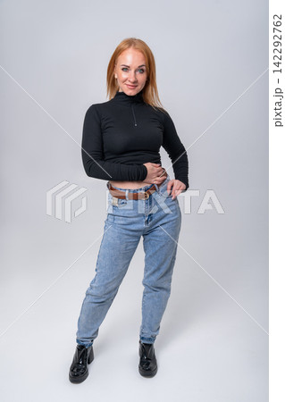 Young woman posing in a studio, wearing a black long sleeved top and light blue jeans, hands resting on her hips, expressing confidence and casual style Young woman posing in a studio, wearing a black long sleeved top and light blue jeans, hands resting on her hips, expressing confidence and casual style 142292762