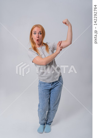 Redheaded woman standing against a grey studio background, playfully flexing her bicep and pointing at it with a surprised, humorous expression conveying strength and fitness 142292764