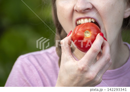 Young woman biting into a fresh organic red tomato, enjoying a healthy snack from the garden with a natural nutritious summer taste 142293341
