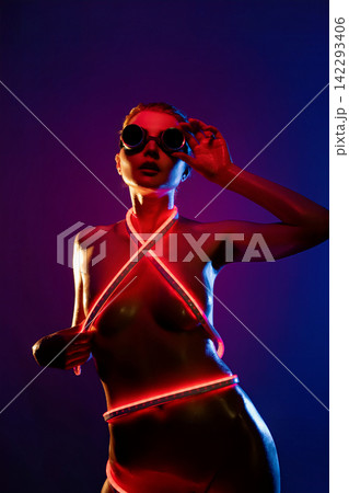Woman in neon red glow with futuristic goggles and glowing straps against dark purple background Woman in neon red glow with futuristic goggles and glowing straps against dark purple background 142293406