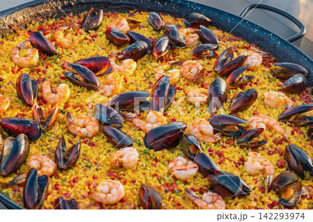 Colorful seafood paella with shrimp, mussels and yellow rice cooked in large traditional pan. Fresh Mediterranean rice dish prepared for outdoor catering and food festival. Colorful seafood paella with shrimp, mussels and yellow rice cooked in large traditional pan. Fresh Mediterranean rice dish prepared for outdoor catering and food festival. 142293974