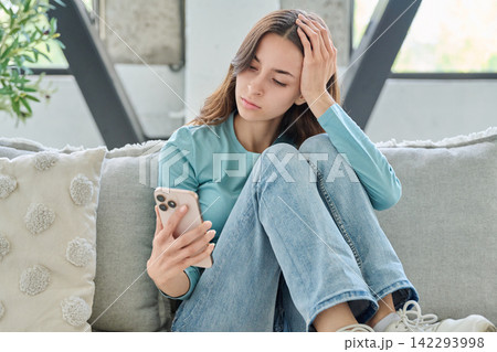 Upset young woman looking at smartphone reading bad news on couch Upset young woman looking at smartphone reading bad news on couch 142293998