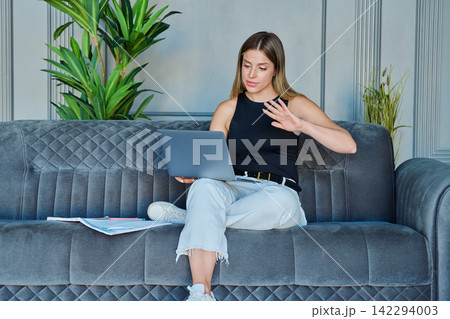 Young woman waving hand during online video call on laptop sitting on sofa at home Young woman waving hand during online video call on laptop sitting on sofa at home 142294003