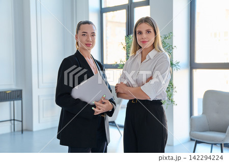 Two businesswomen standing together looking at camera holding laptop in modern bright office Two businesswomen standing together looking at camera holding laptop in modern bright office 142294224