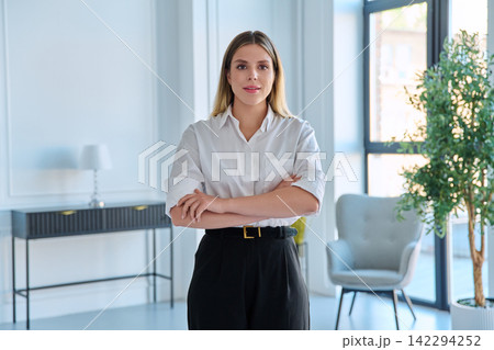 Smiling young business woman standing with arms crossed in modern light office Smiling young business woman standing with arms crossed in modern light office 142294252