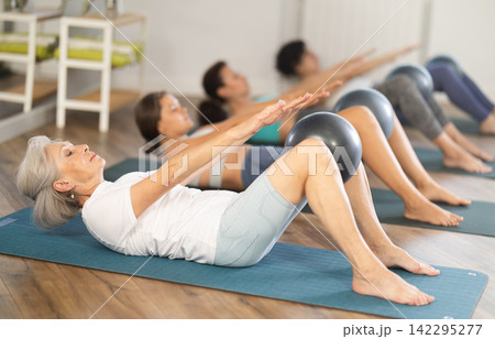 Elderly woman performing Half Roll Back with ball during pilates class 142295277