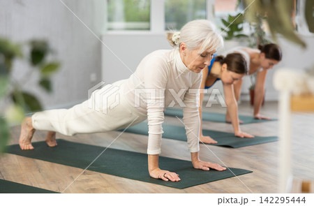 Focused older woman doing yoga poses with other women in yoga studio Focused older woman doing yoga poses with other women in yoga studio 142295444
