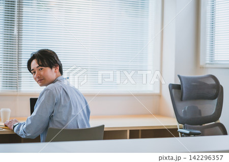 デスクで振り向いて笑顔を見せる男性ビジネスパーソンのオフィスシーン 142296357