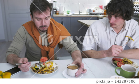 A family of young gay men quarrels over breakfast. Two men are eating at a table set with plates of food and cups of coffee A family of young gay men quarrels over breakfast. Two men are eating at a table set with plates of food and cups of coffee 142297500