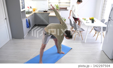 A guy exercises while his boyfriend has breakfast. A man takes a yoga pose on a blue rug in the kitchen. The ordinary life of gays A guy exercises while his boyfriend has breakfast. A man takes a yoga pose on a blue rug in the kitchen. The ordinary life of gays 142297504