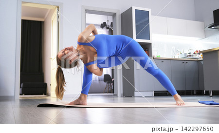 A sexy female yogi stands in a complex asana on a yoga mat. She practices oriental practices in her living room against the background of the kitchen 142297650